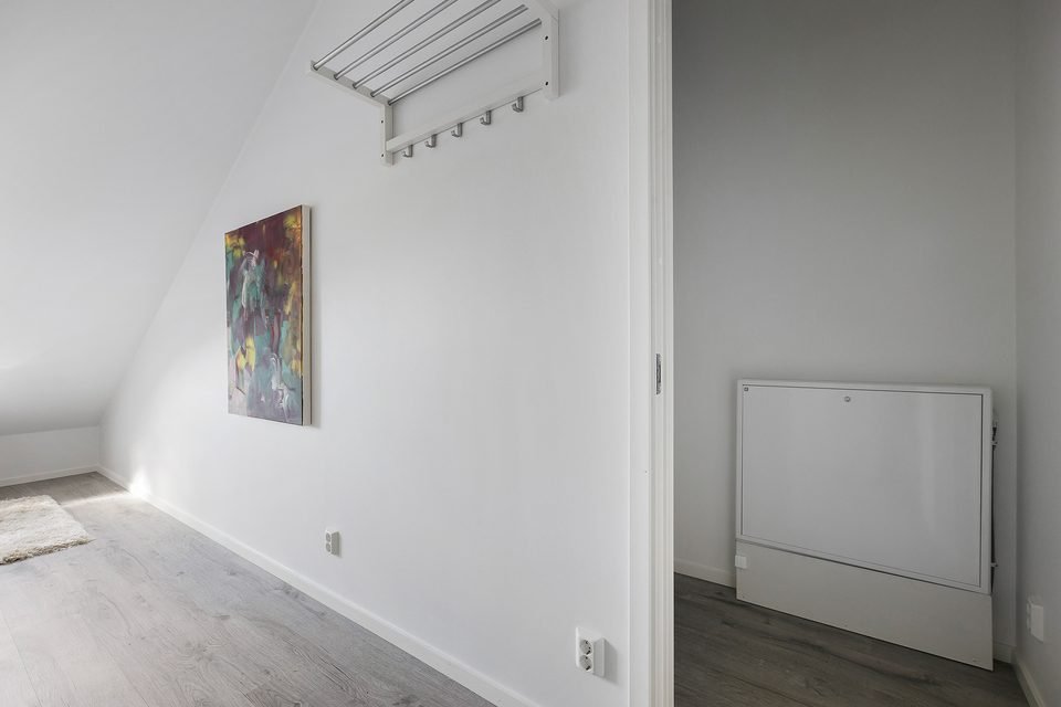 Vägg i hallen med tavla, hatthylla och en vit radiator under snedtaket.