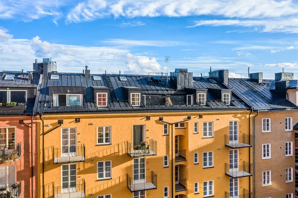 Husets gula fasad och plåttak sedda från luften, med parkträd och höga hus i bakgrunden.