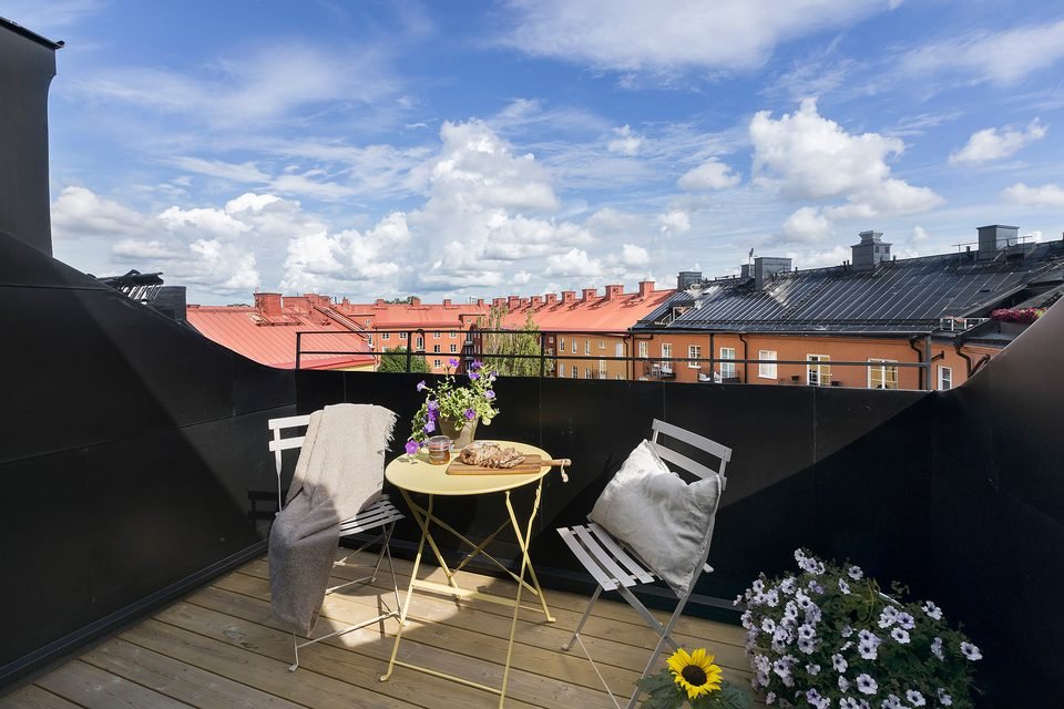 Takterrass med trädäck, bord och två stolar, blommor i kruka och utsikt över omgivande hustak.