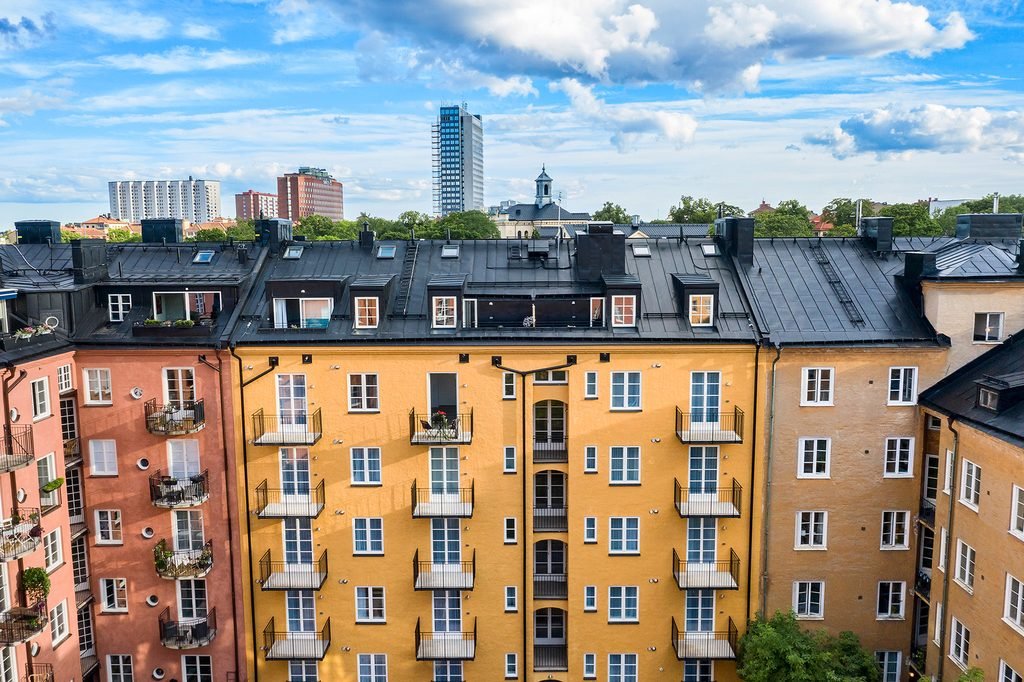 Fastigheten sedd snett ovanifrån. Gul putsad fasad med balkonger, plåttak med takkupor och Stockholms hustak i bakgrunden.